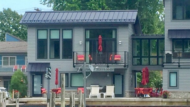 Un bâtiment situé au bord de l’eau, doté d’un quai, de nombreuses fenêtres et d’un espace extérieur aménagé avec des parasols rouges.