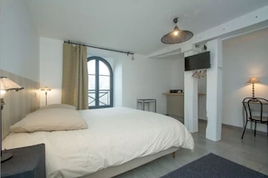 Een slaapkamer met een groot bed, een raam met gordijnen, een aan de muur bevestigde televisie en een kleine tafel met een lamp.
