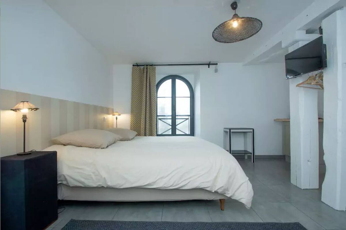 Un dormitorio con cama, lámparas de noche, una mesita, un televisor montado en la pared y una ventana con cortinas.