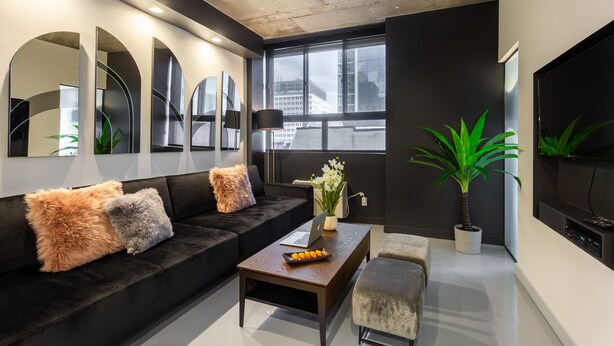 A modern living room with a black sofa, a wooden coffee table, a TV, and a large window with a city view. - Ambient Loft with All Amenities (Montréal)