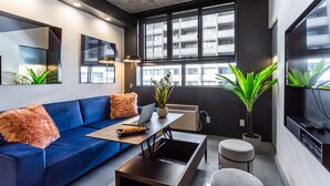 Un salon moderne avec un canapé bleu, une table basse, une petite table avec un ordinateur portable et une grande fenêtre donnant sur la ville.