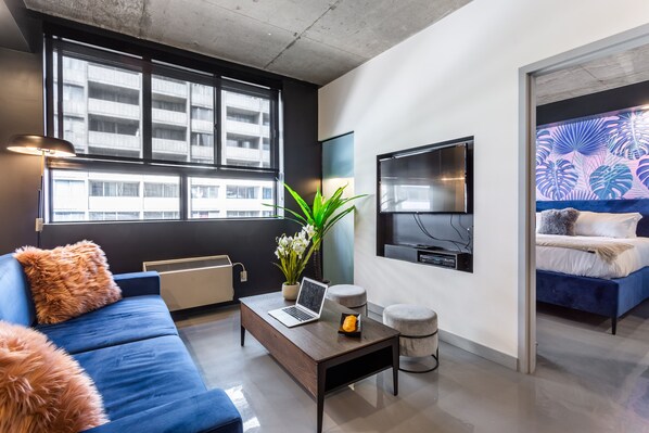 Un salón moderno con un sofá azul, una mesa de centro con una computadora portátil, una planta en maceta y un ventanal con vista a un edificio.