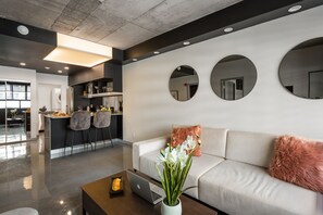 Un salon moderne avec un canapé, une table basse et un ordinateur portable. On aperçoit à l’arrière-plan un coin cuisine avec une table à manger et des chaises.