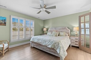 Une chambre à coucher avec un lit, un ventilateur de plafond, une fenêtre avec des volets, une table de chevet, une lampe, une chaise en osier et une porte.