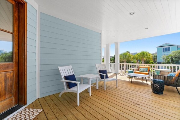 A covered porch with white chairs, a table, and a black chair with orange cushions.