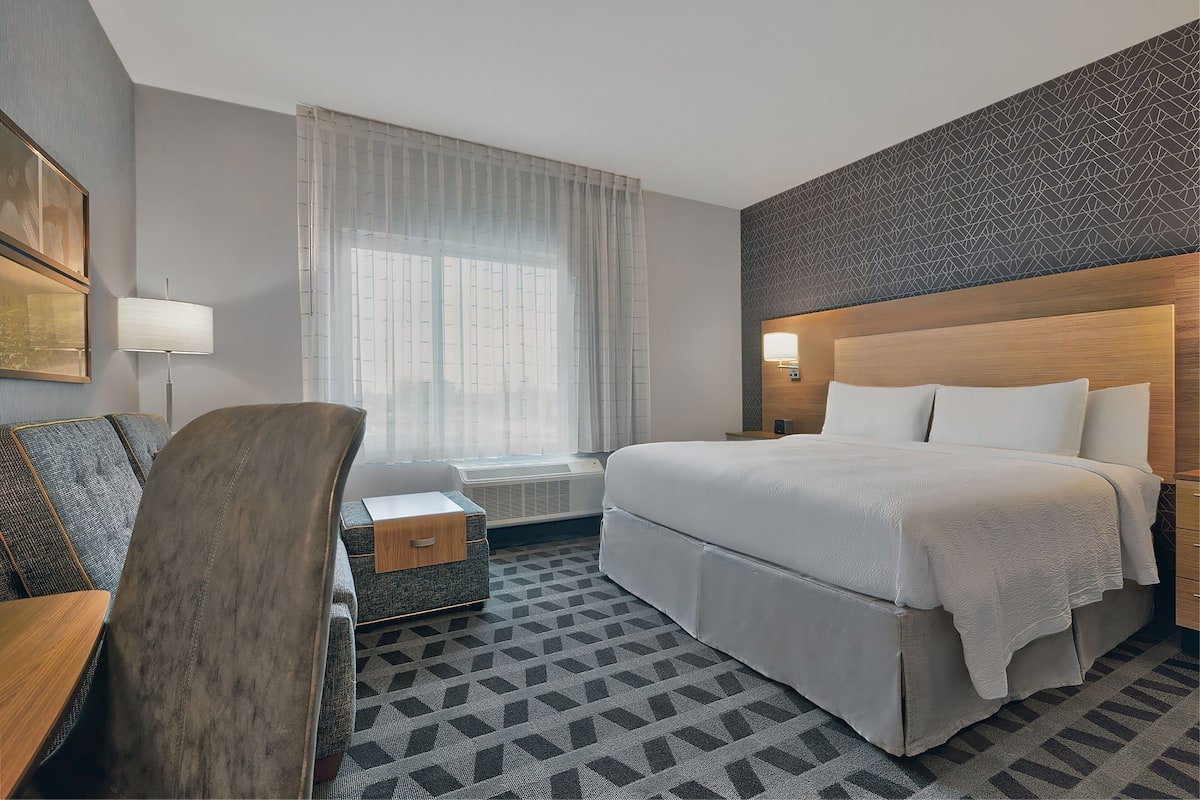 A hotel room with a bed, a desk, a chair, a window with curtains, and a patterned carpet.