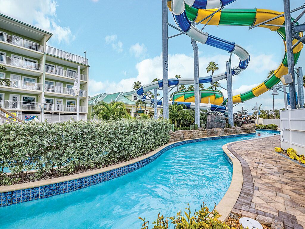 A pool area with a water slide, a multi-story building with balconies, and a clear blue sky.