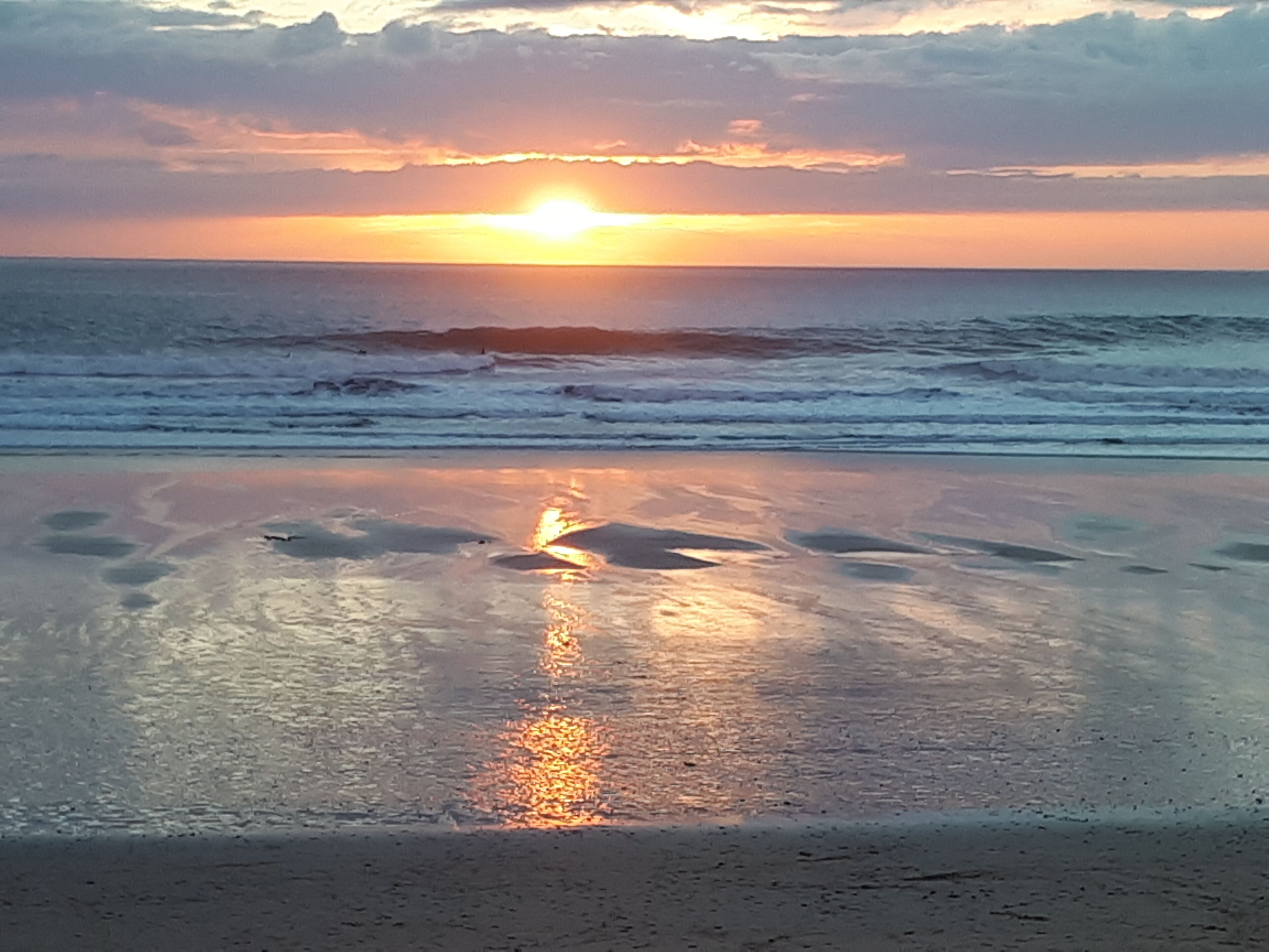 A beach scene with the sun setting over the ocean, reflecting on the wet sand.