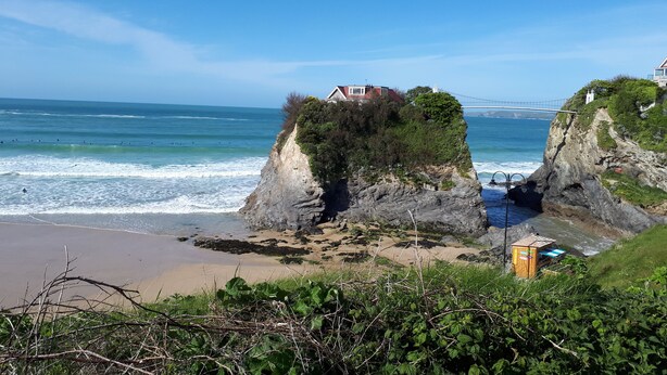 Paesaggio costiero con una casa a picco sulla scogliera, una spiaggia e un ponte sullo sfondo.