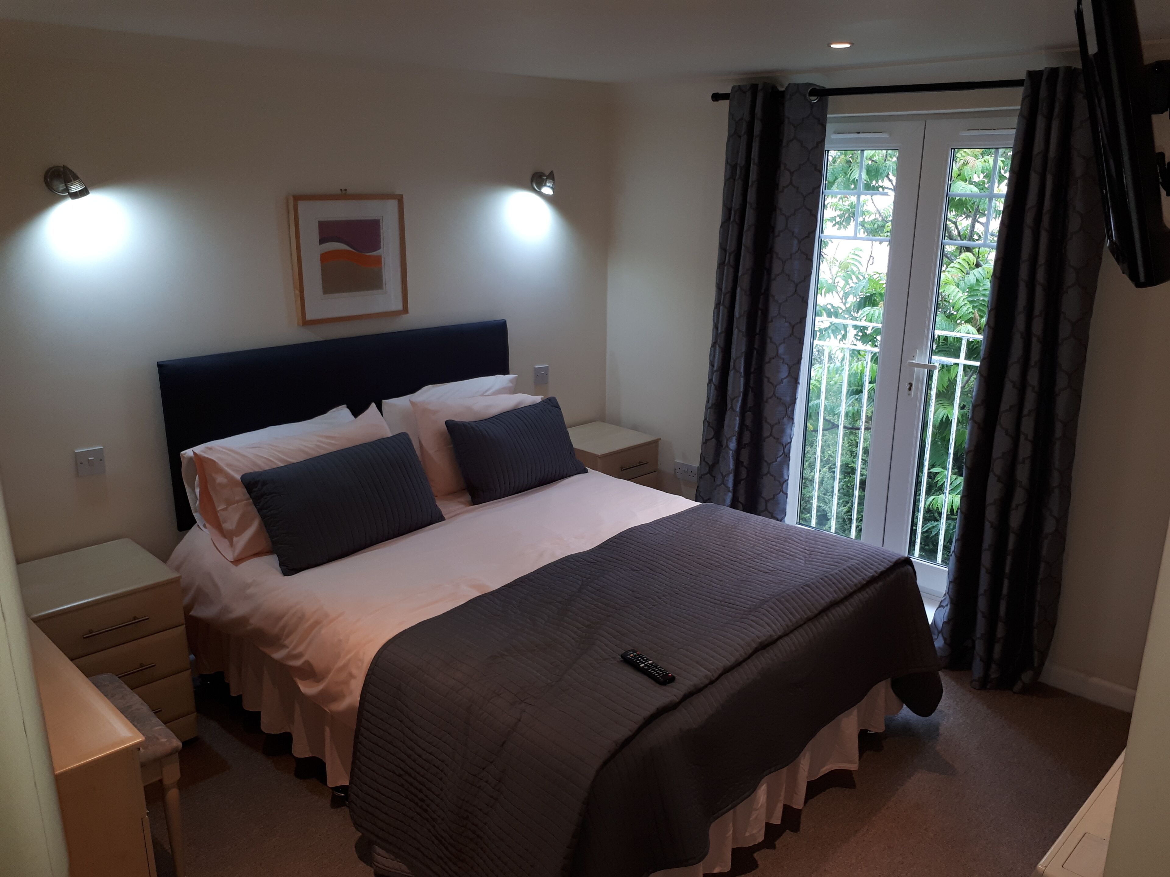 Une chambre d’hôtel avec un lit, des tables de chevet, une commode, un tableau encadré au mur et une fenêtre avec des rideaux.