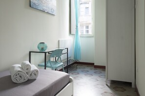 A neatly made bed with rolled white towels, a small desk with a blue chair, a window with a blue curtain, and patterned floor tiles.