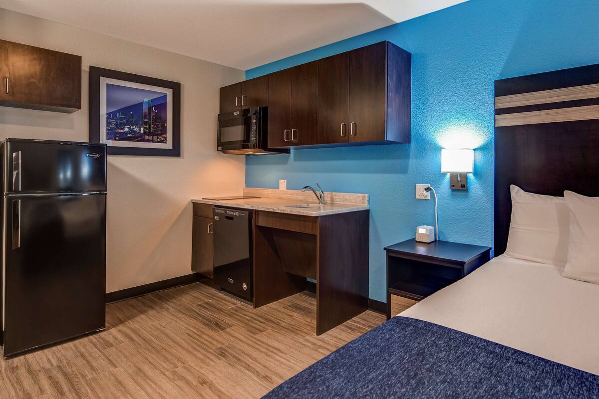A compact hotel room with a bed, a small kitchenette, a microwave, a refrigerator, and a nightstand.