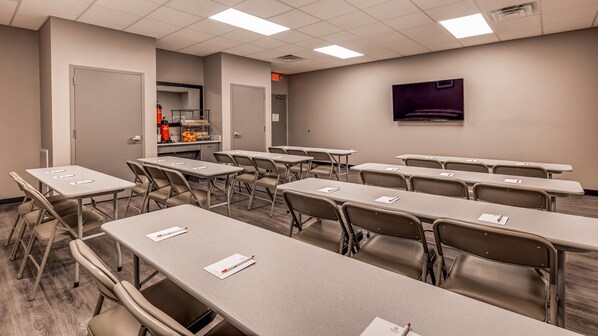 Uma sala de conferências com várias mesas e cadeiras, um espaço de kitchenette e uma tela grande na parede.