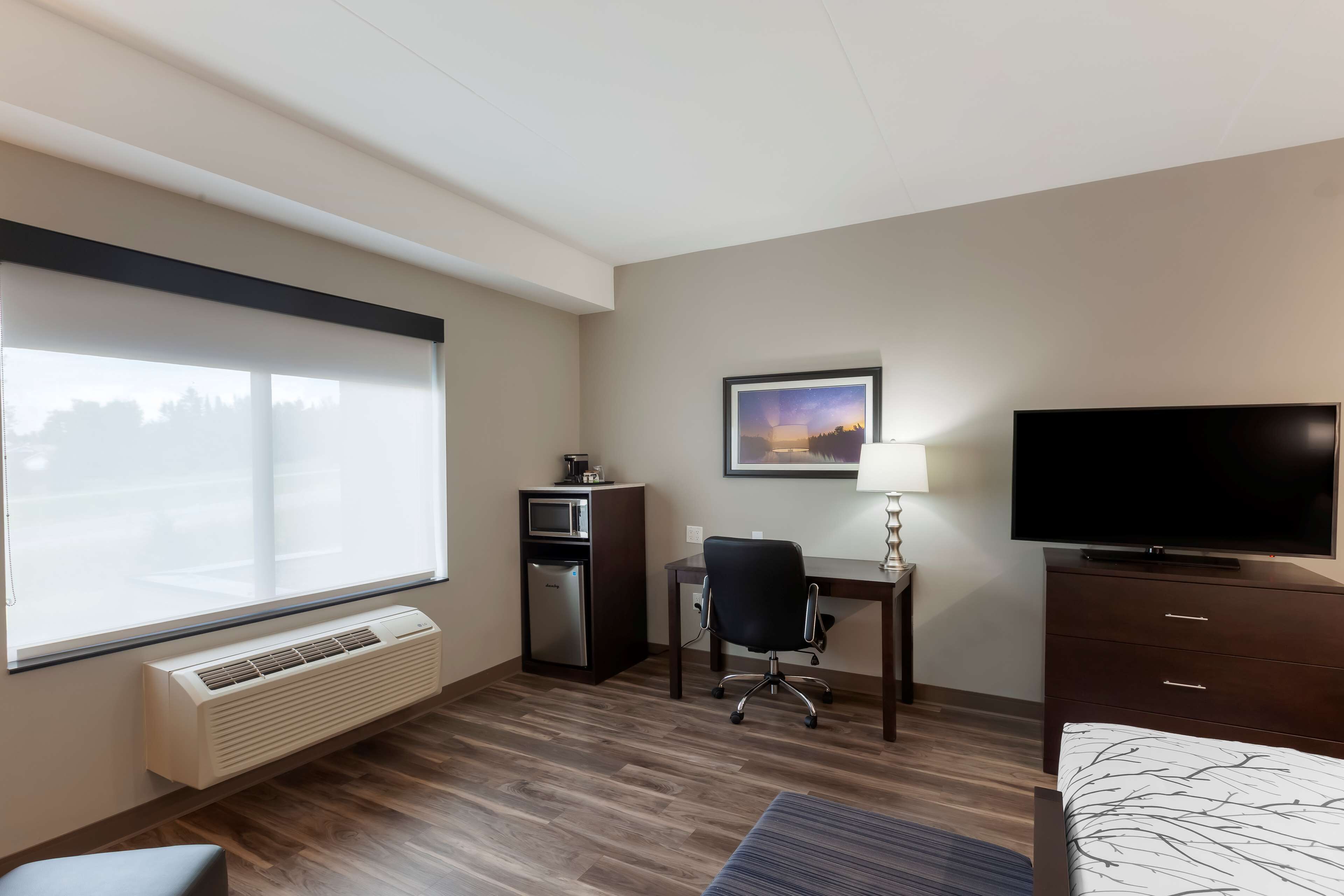 Habitación de hotel con un ventanal grande, un escritorio con silla, televisión, cama y pequeña cocina.