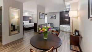 Habitación de hotel con cama, zona de estar con sofá y sillones, mesa de comedor con sillas, baño con bañera y un espejo grande en la pared.