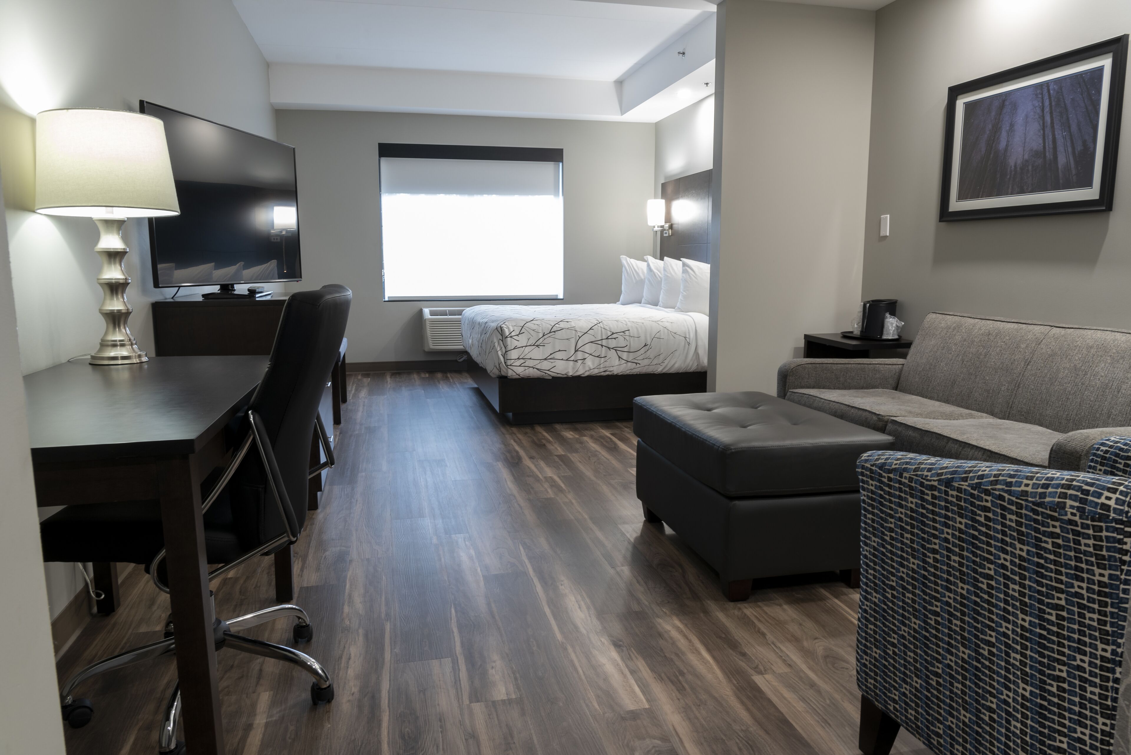 A modern hotel room with a flat-screen TV, a desk with a lamp, a sofa, an ottoman, and a large window with blinds.