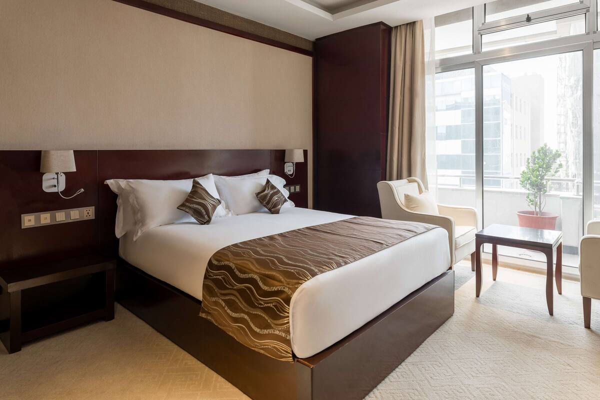 A hotel room with a large bed, bedside tables, a chair, a small table, and a window with a view of buildings.