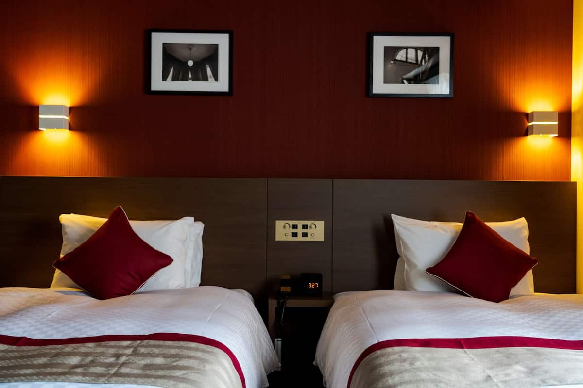 A hotel room with two beds, each with white bedding and red pillows, a nightstand with a clock, and two framed pictures on the wall.