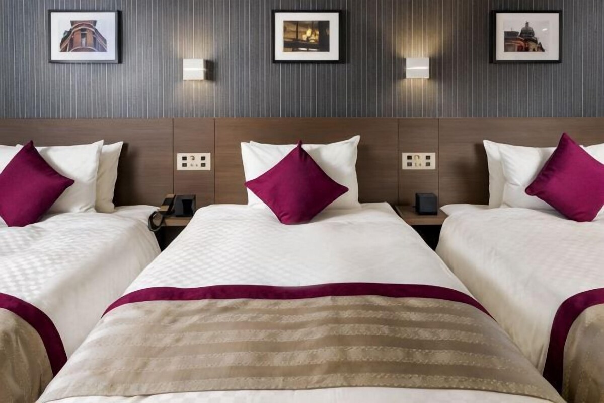 A hotel room with three beds, each with a pillow and a blanket. There are wall-mounted lights and a framed picture above the beds.