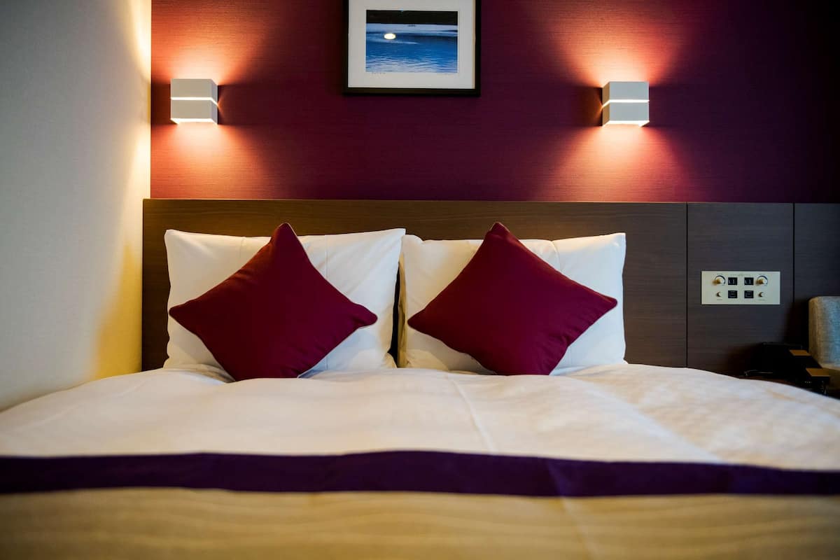 A neatly made bed with white and maroon pillows, a framed picture on the wall, and two wall-mounted light fixtures.