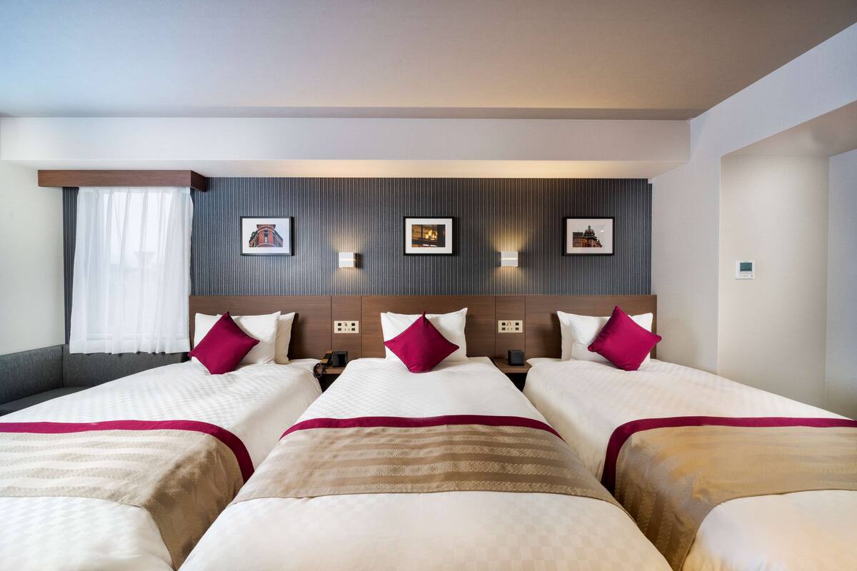 A hotel room with four beds, a large window with sheer curtains, and wall-mounted light fixtures.