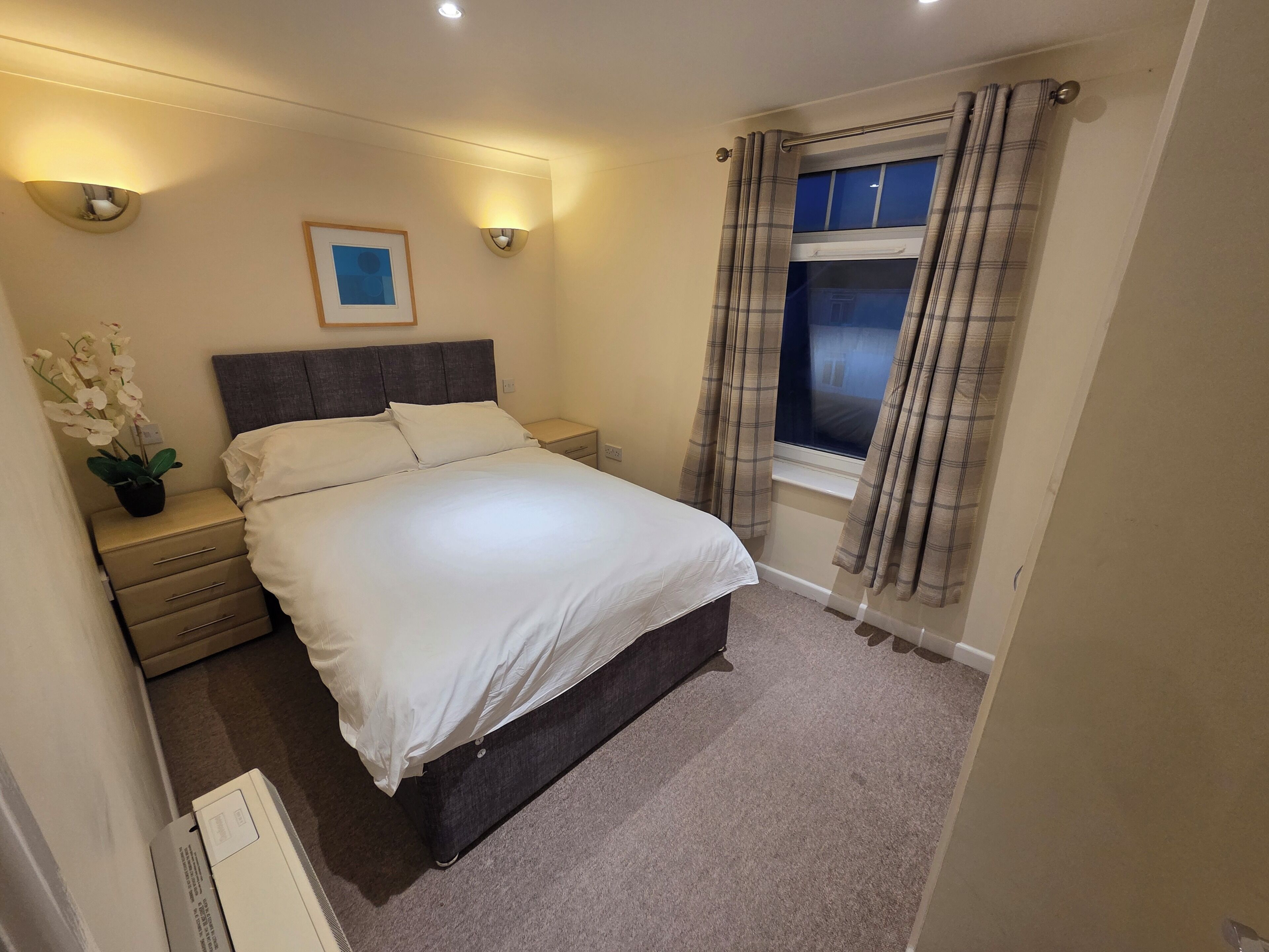 A bedroom with a bed, bedside tables, a nightstand, a window with curtains, and a wall-mounted light fixture.