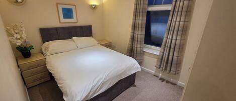 A bedroom with a bed, bedside tables, a nightstand, a window with curtains, and a wall-mounted light fixture.