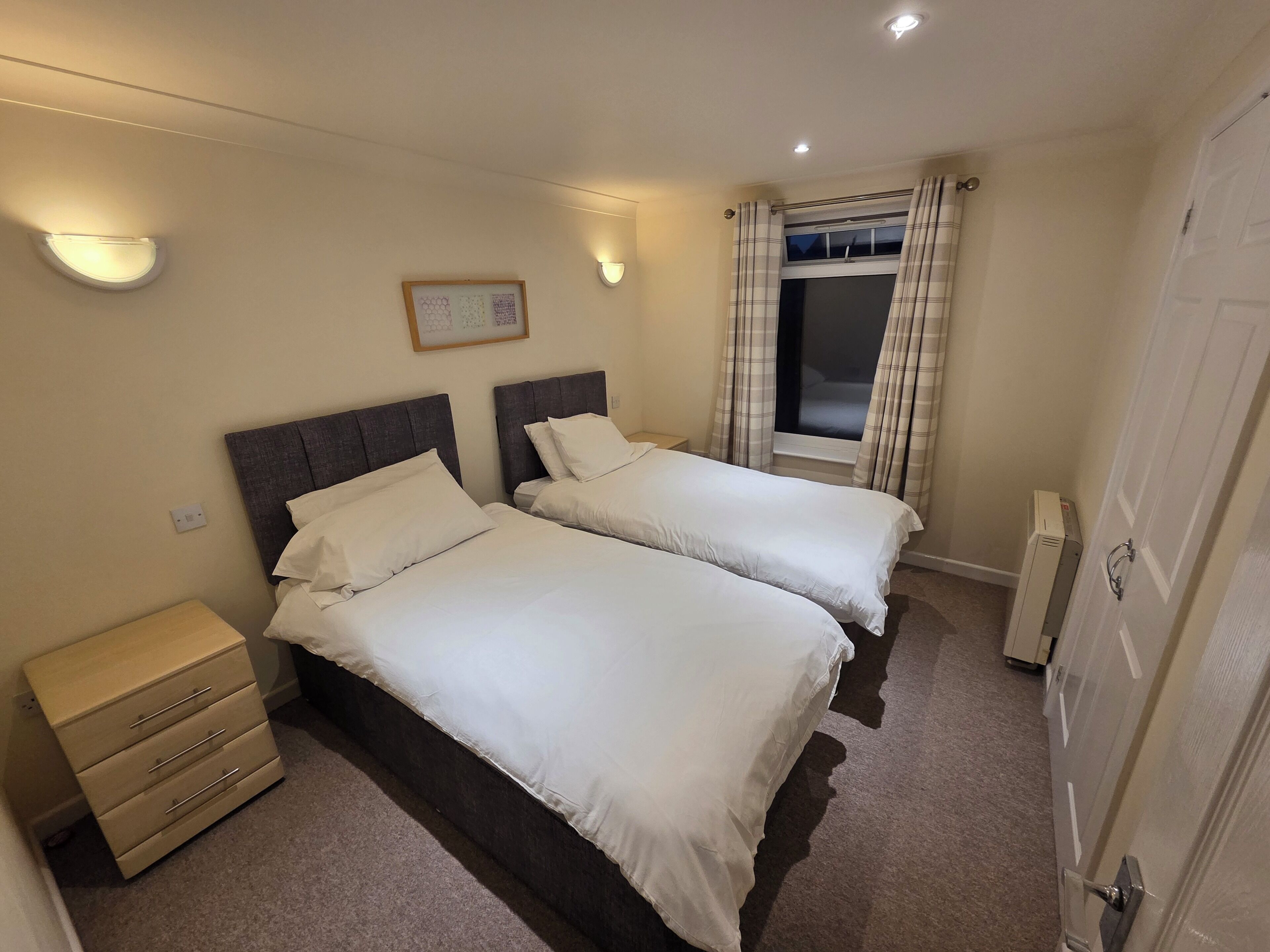 A hotel room with two beds, a bedside table, a dresser, a window with curtains, and a wall-mounted picture.