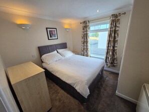 A bedroom with a bed, a bedside table, a wall-mounted light, a framed picture, and a window with curtains.