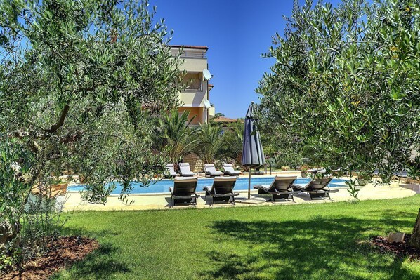 A pool area with lounge chairs and umbrellas, surrounded by greenery and a building in the background. - The sandy beach is only a few minutes' walk away (Medulin)