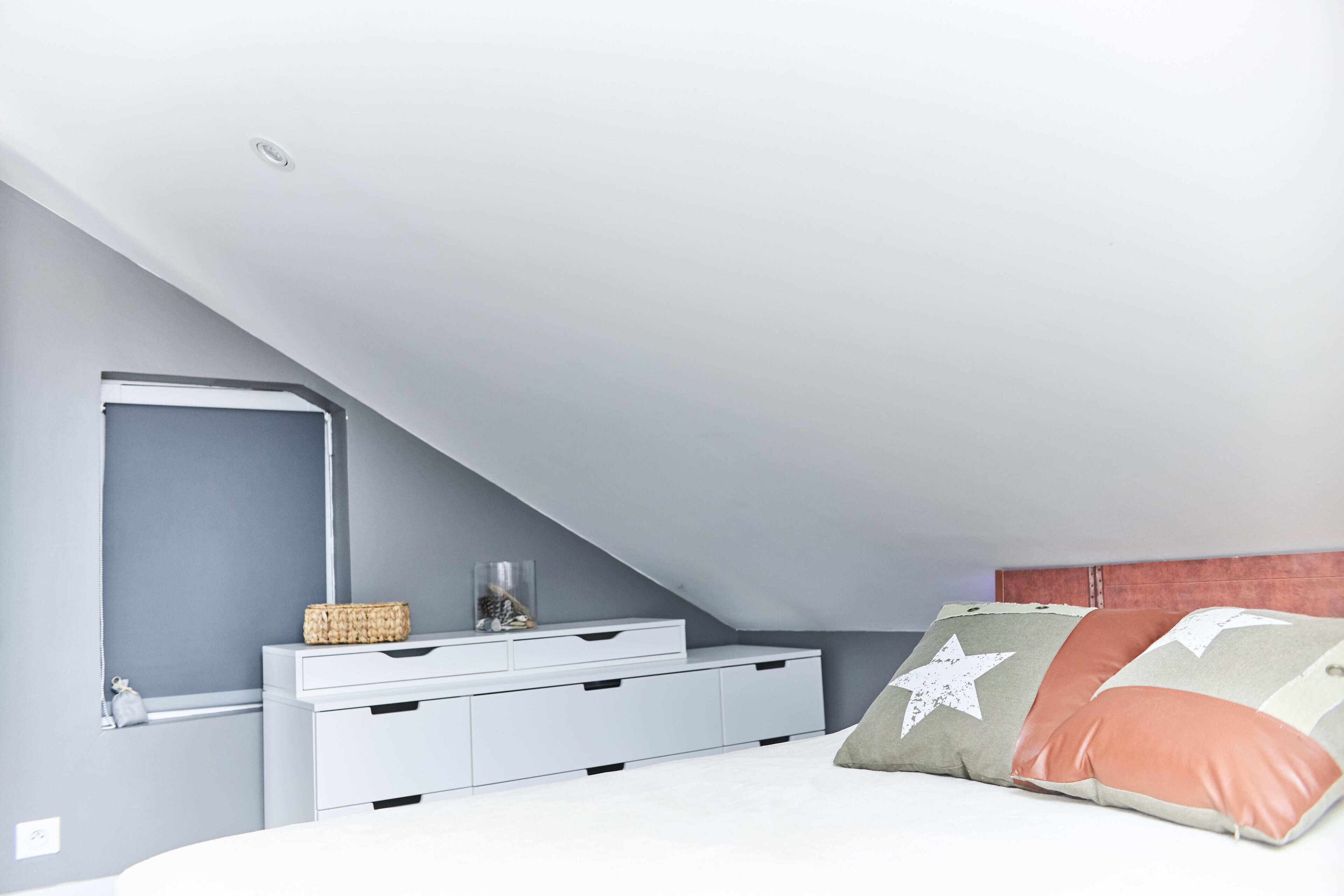 Une chambre avec un plafond en pente, un lit blanc et une table de chevet avec des tiroirs.