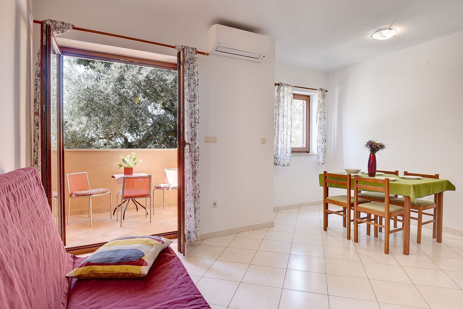 A room with a balcony, a dining area with a table and chairs, and a sofa with a colorful cushion.