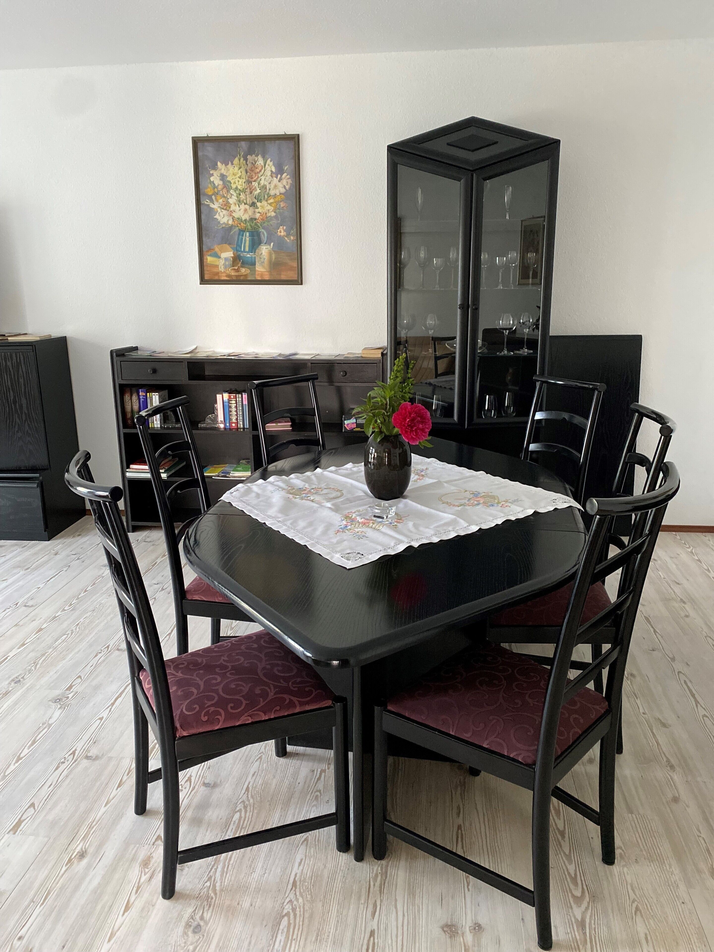 A dining area with a black table, red cushioned chairs, a vase with flowers, and a bookshelf in the background.