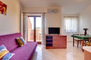Una habitación con sofá morado, un mueble de televisión de madera, una mesa de comedor con sillas y un ventanal corredizo que da a un espacio exterior.