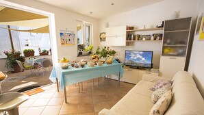 Una sala de estar con un sofá, una mesa de comedor con mantel azul, un televisor y varias plantas en macetas.