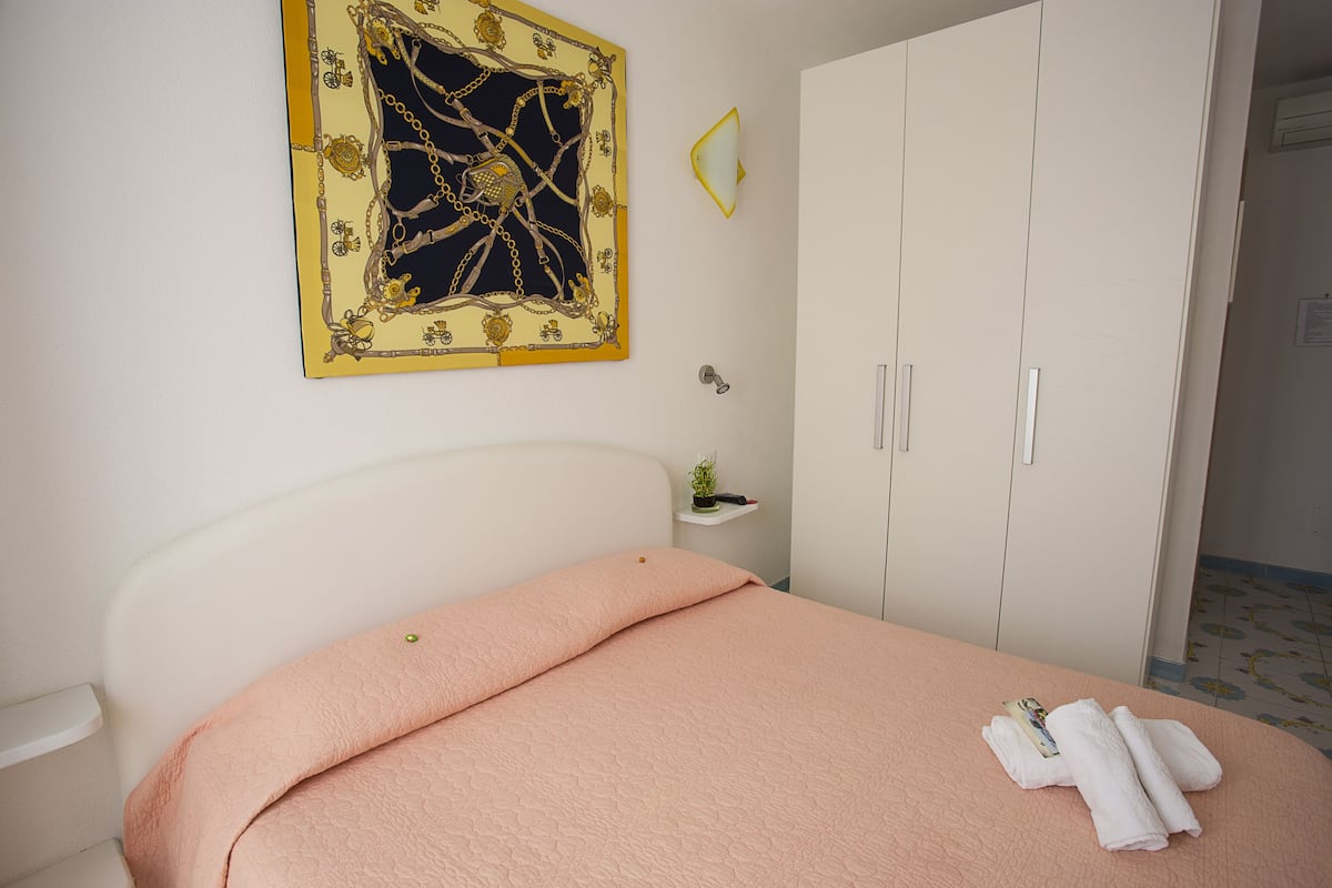 A bedroom with a bed, a wardrobe, a small shelf with a plant, and a framed artwork on the wall.