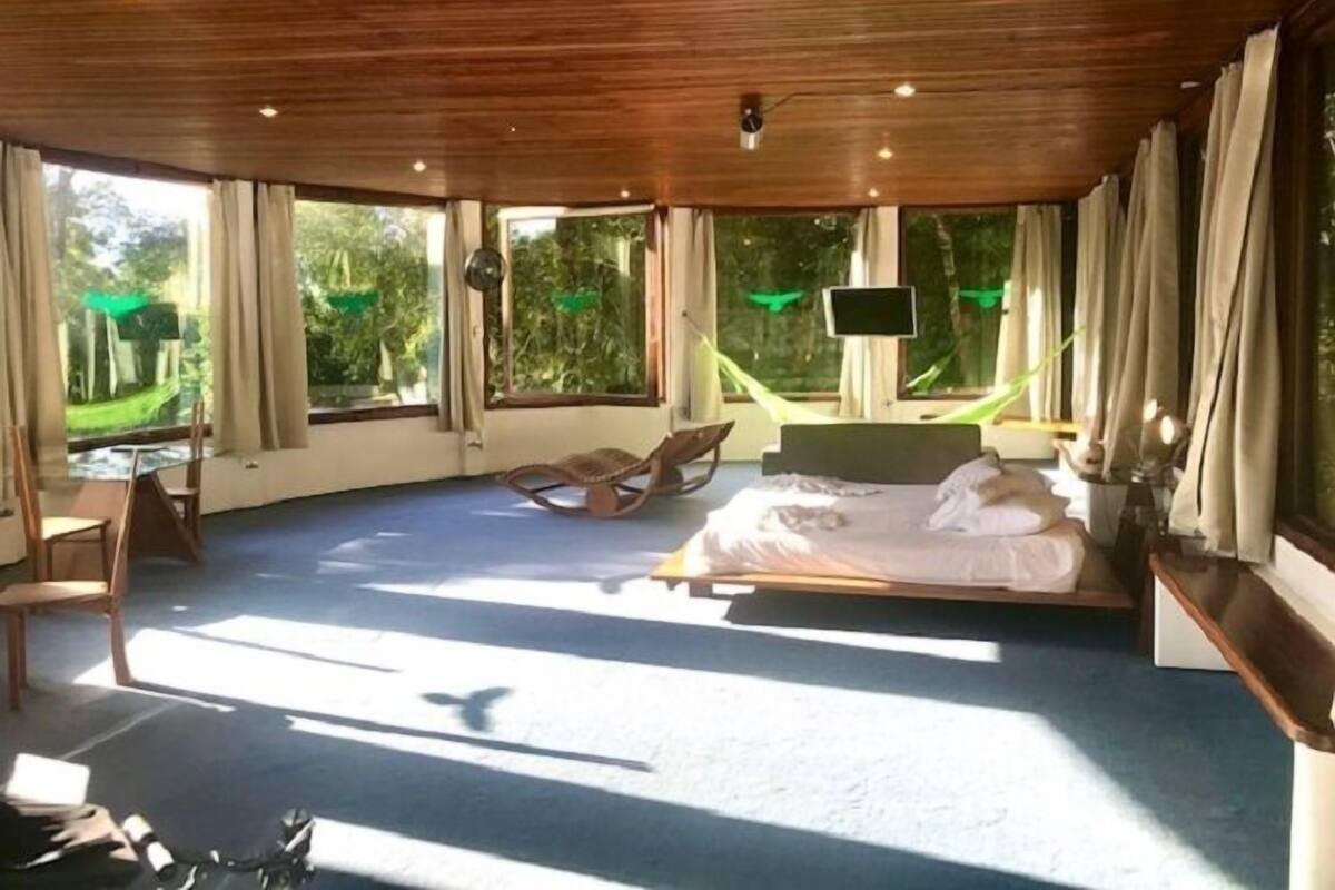 Une chambre spacieuse avec un grand lit, un bureau et une chaise. De grandes fenêtres sont agrémentées de rideaux, et on y trouve également un hamac et un téléviseur posé sur un meuble.