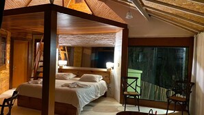 A cozy bedroom with a wooden ceiling, a large bed, a sitting area with a bathtub, and a view through a large window. - Jardim Suspenso da Babilônia (Santo Antonio do Pinhal)