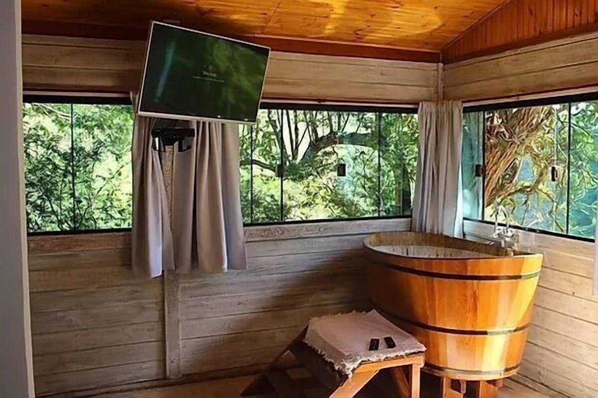 Une cabane en bois avec une baignoire en bois, un téléviseur fixé au mur et de grandes fenêtres donnant sur la forêt.