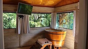 A wooden cabin with a wooden bathtub, a television mounted on the wall, and large windows offering a view of trees. - Jardim Suspenso da Babilônia (Santo Antonio do Pinhal)