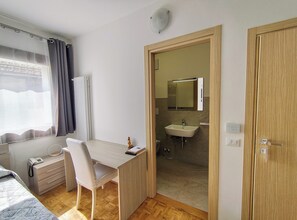 A room with a wooden desk, a chair, a bedside table with a lamp, a window with curtains, a radiator, and a door leading to a bathroom. - Hotel da Marco (Vigo di Cadore)