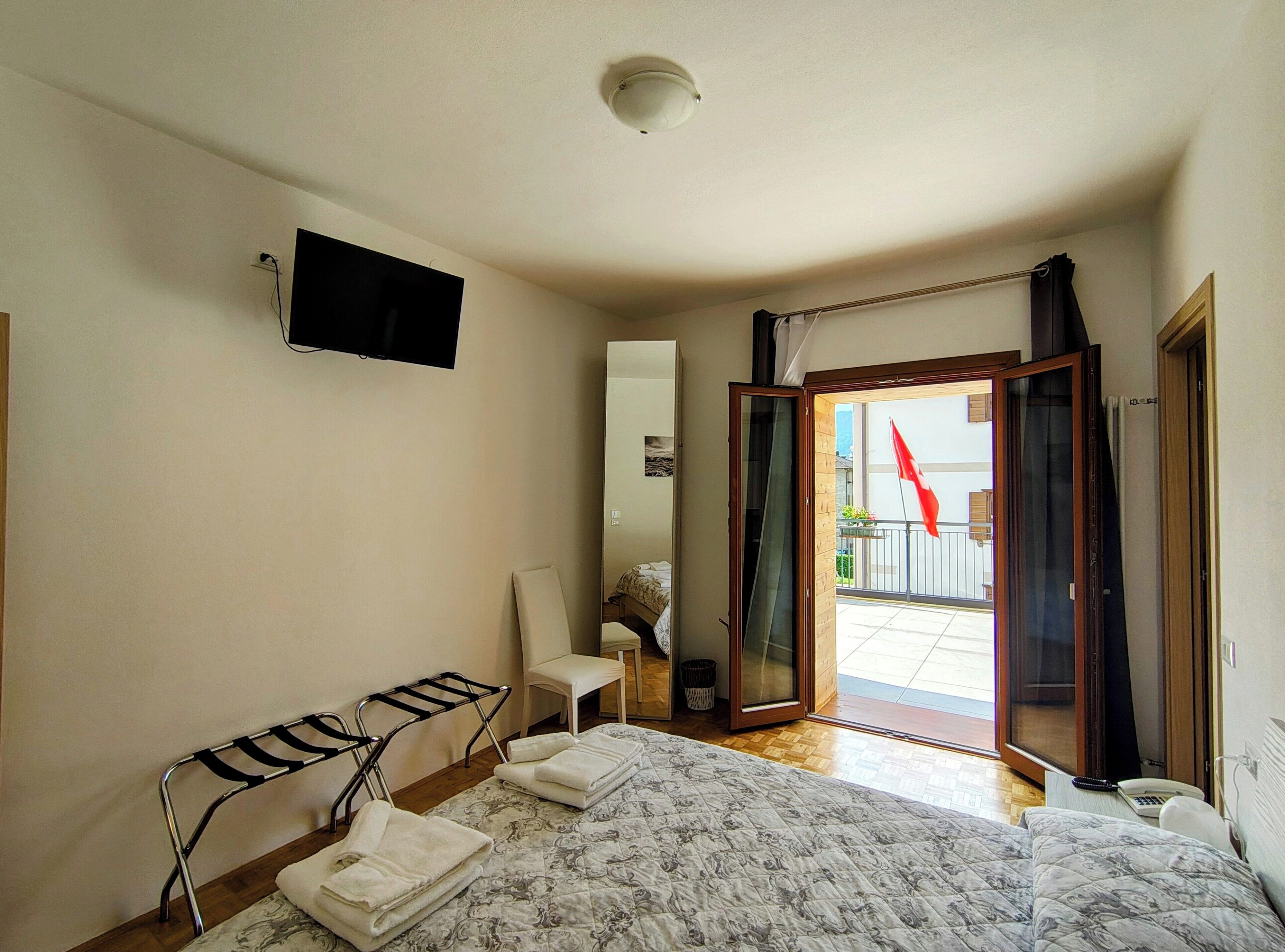 Habitación de hotel con cama, una silla, televisión y balcón con una bandera roja, además de una puerta corrediza.