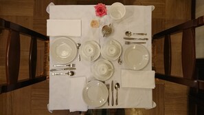 A dining table set with white plates, silverware, and a teapot, with a rose and a small decorative item on the tablecloth.