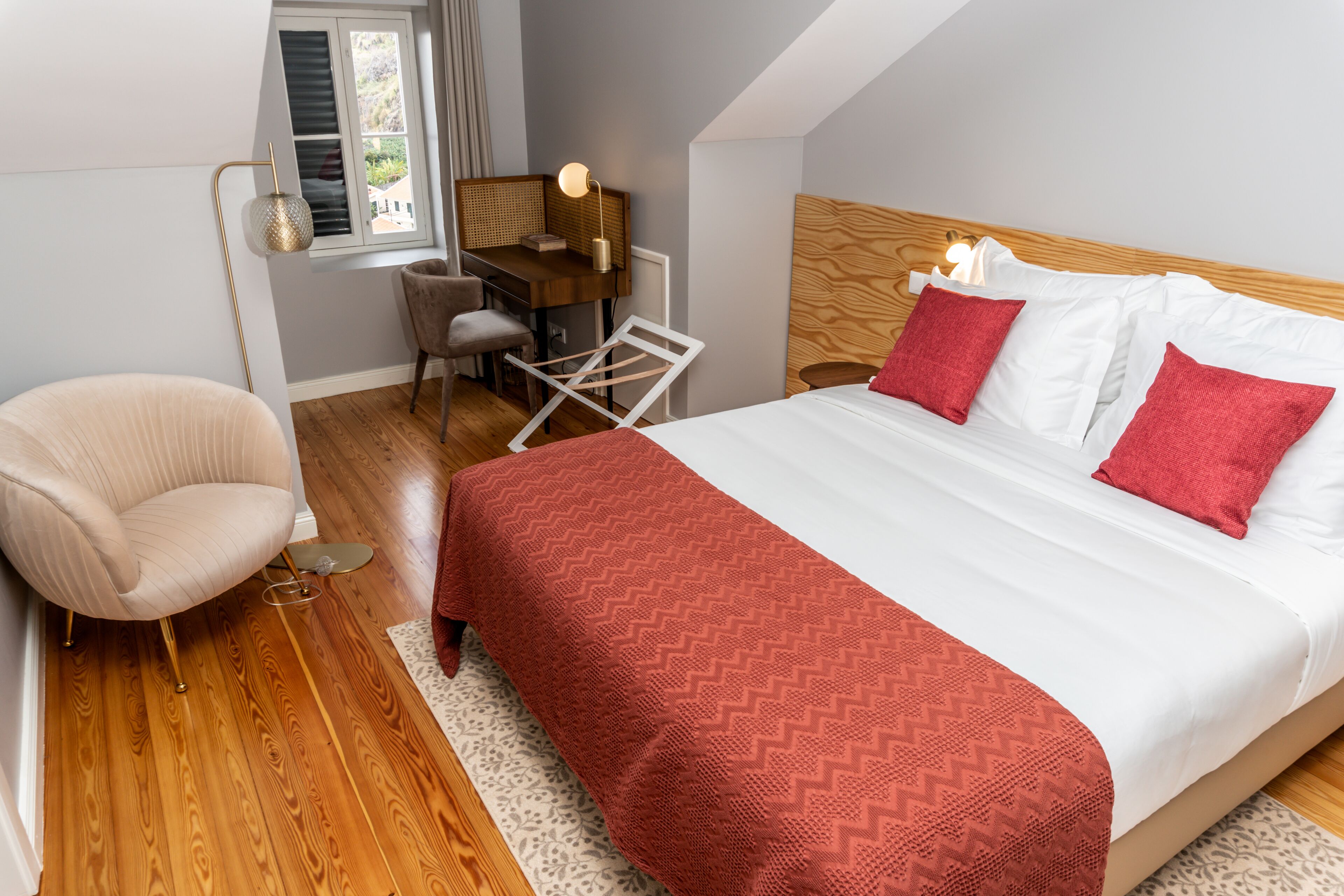 A bedroom with a wooden floor, a bed with white and red bedding, a desk with a chair, and a lamp.