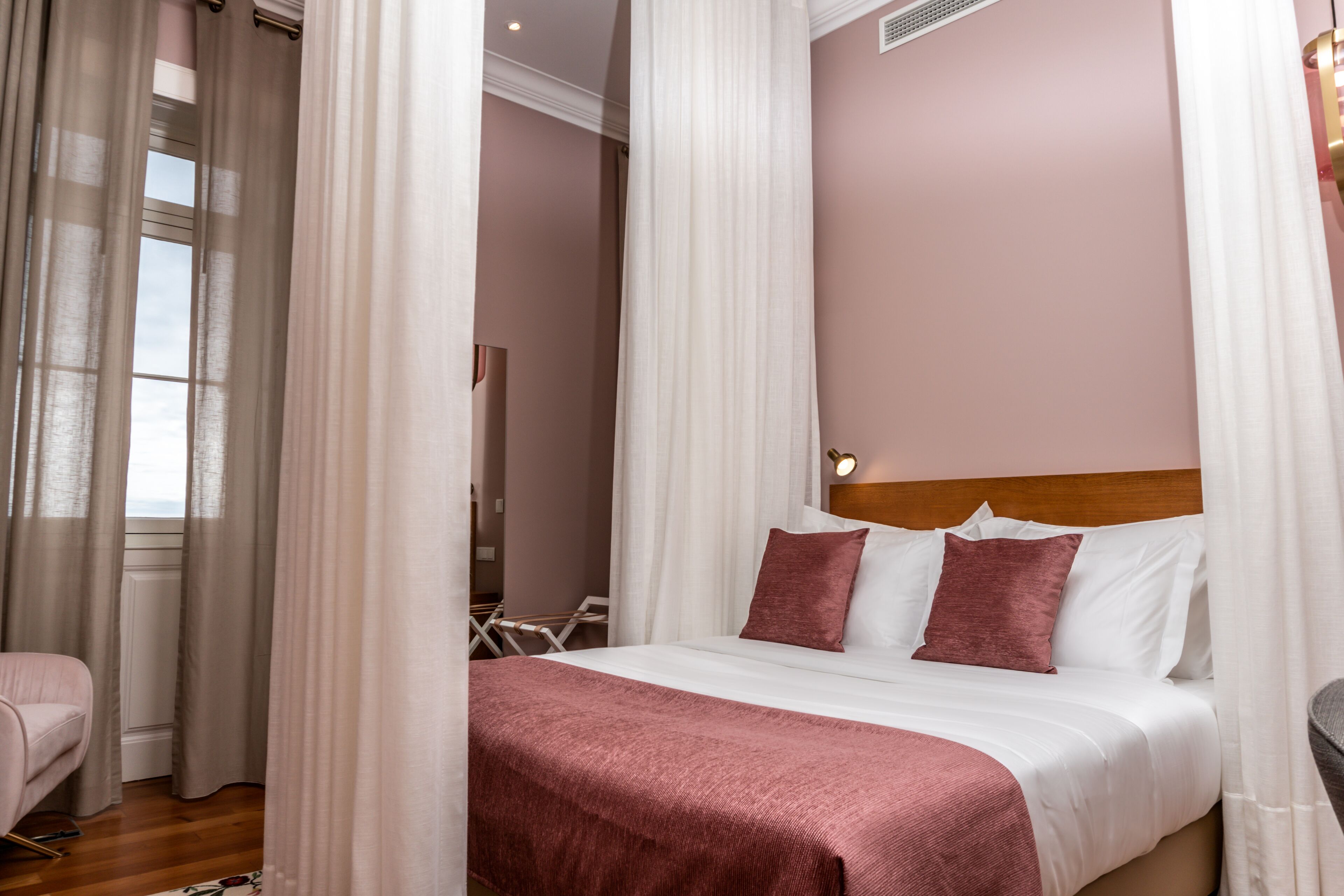 Una cama bien hecha con una colcha rosa y almohadas, un cabecero de madera y cortinas translúcidas ligeramente corridas.