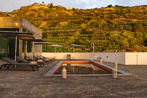 Una piscina en la azotea con sillones de descanso y sombrillas, rodeada de un jardín ajardinado y una ladera.
