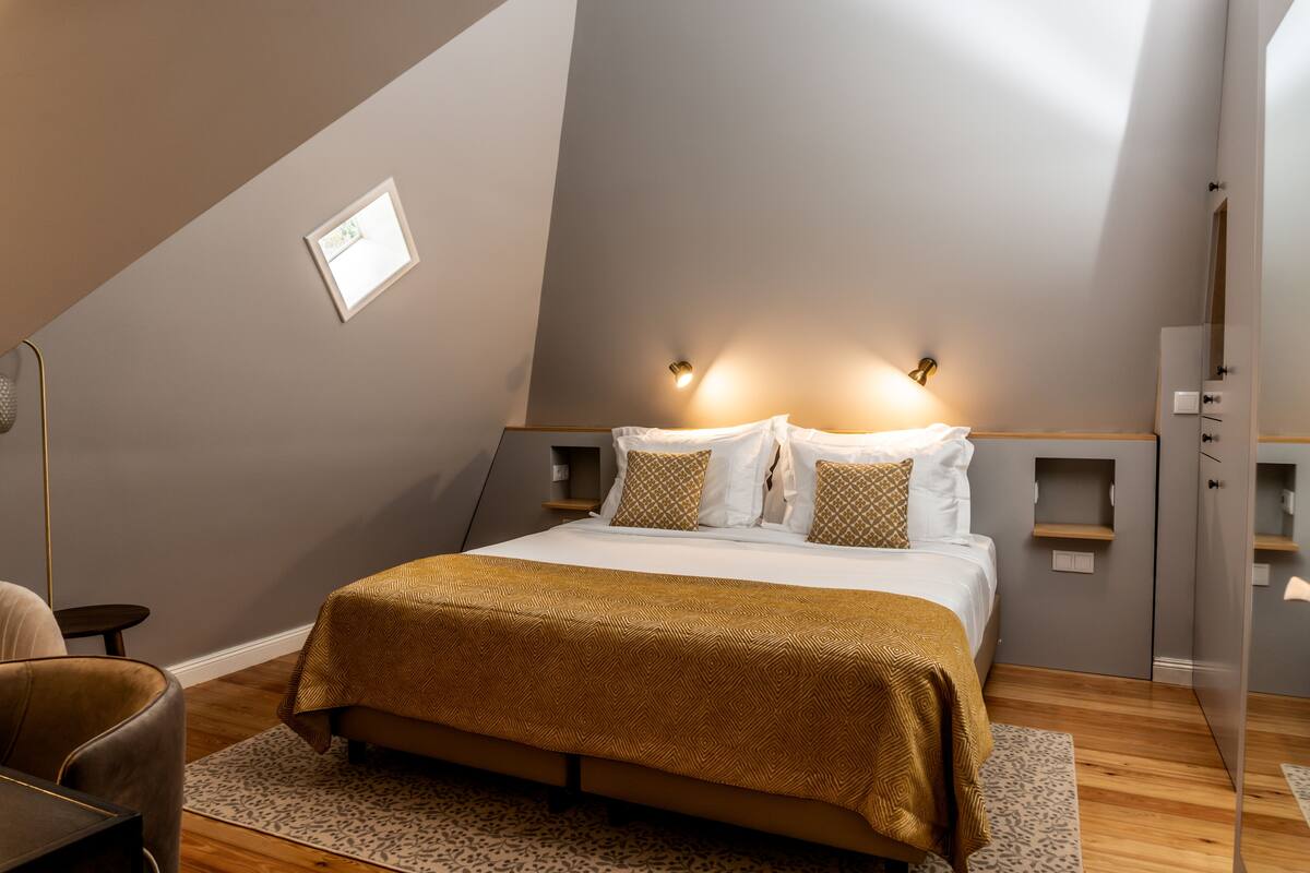 A modern bedroom with a sloped ceiling, a bed with a patterned bedspread, bedside lamps, and a small bedside table.