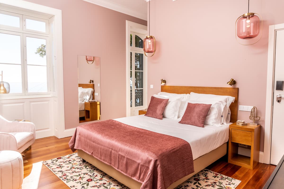 A bedroom with a large bed, a wooden nightstand, a mirror, and a window with a view of trees.