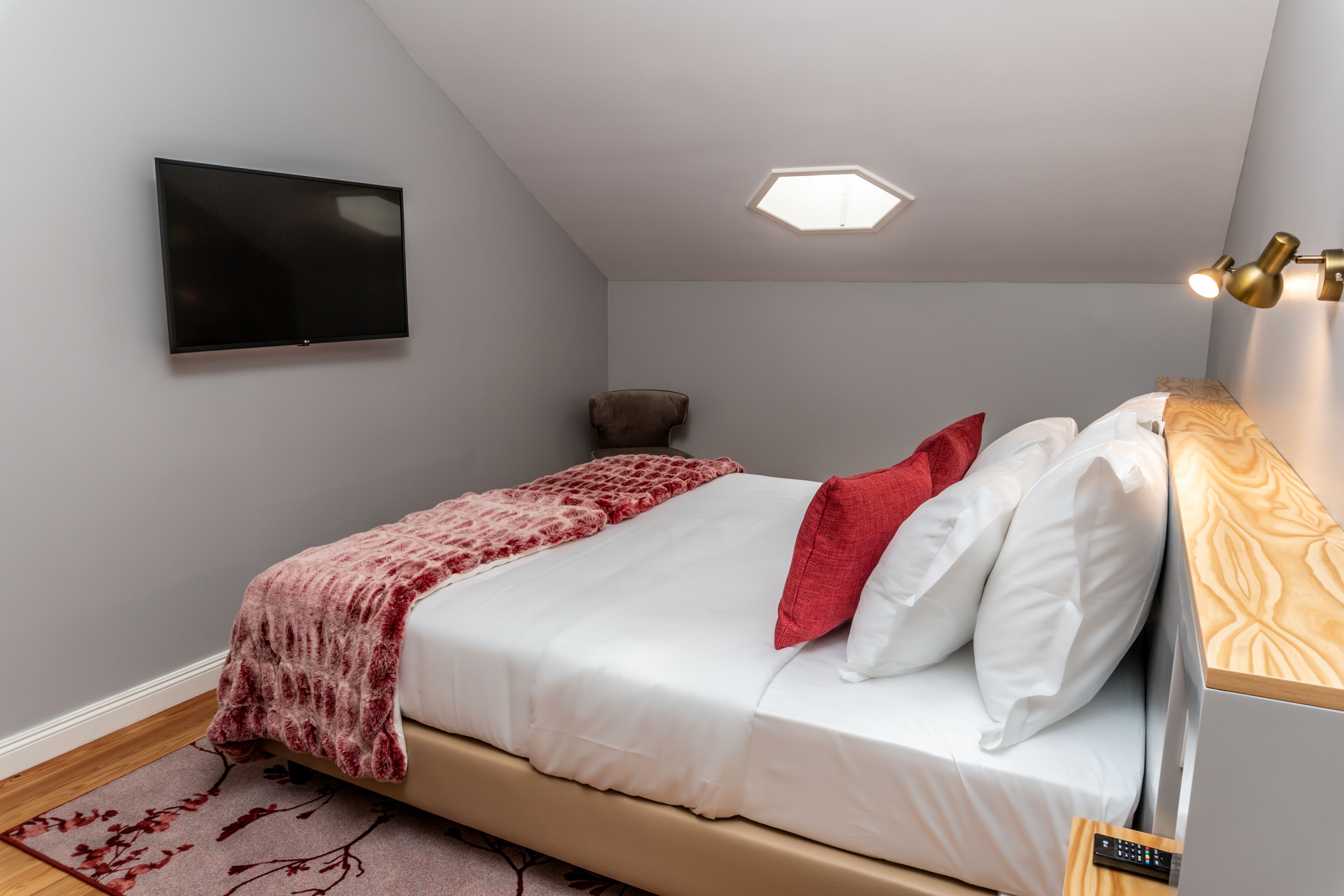 Een slaapkamer met een bed, een wandtelevisie, een plafondlamp en een houten nachtkastje met een afstandsbediening.