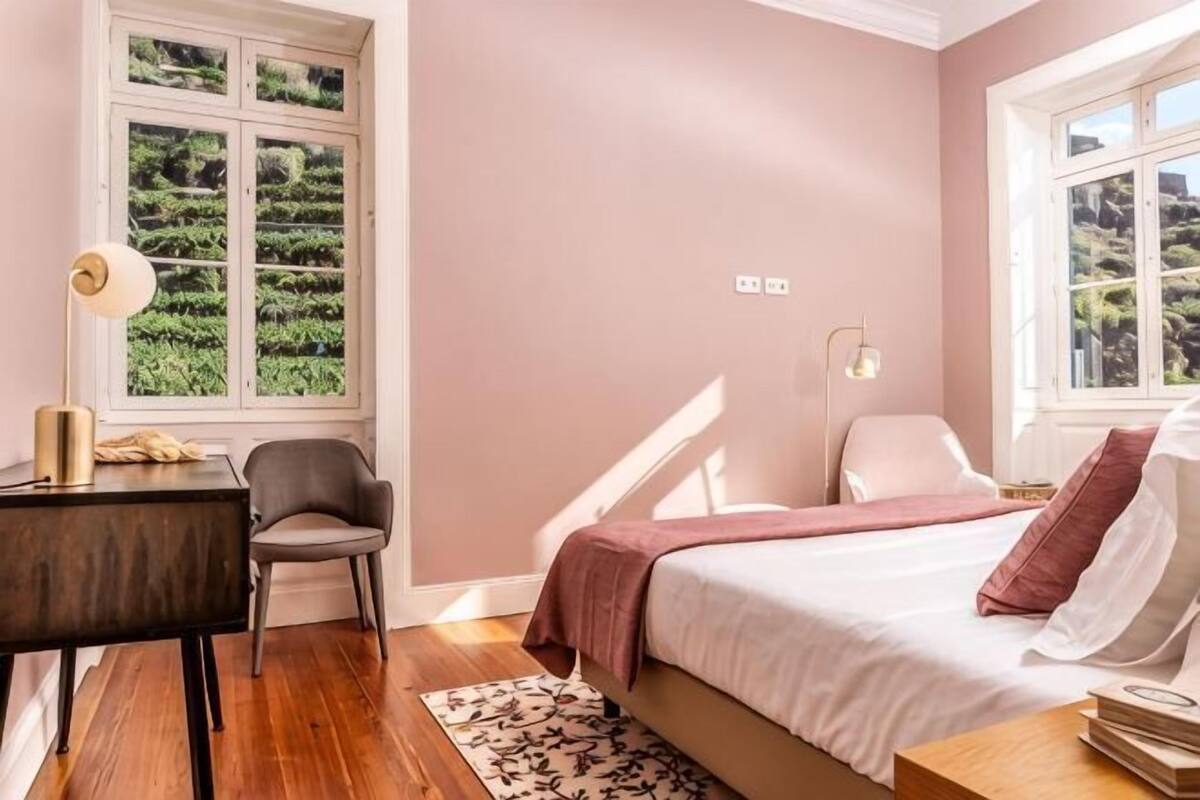 A bedroom with a bed, bedside tables, a desk, a chair, and a window with a view of greenery.